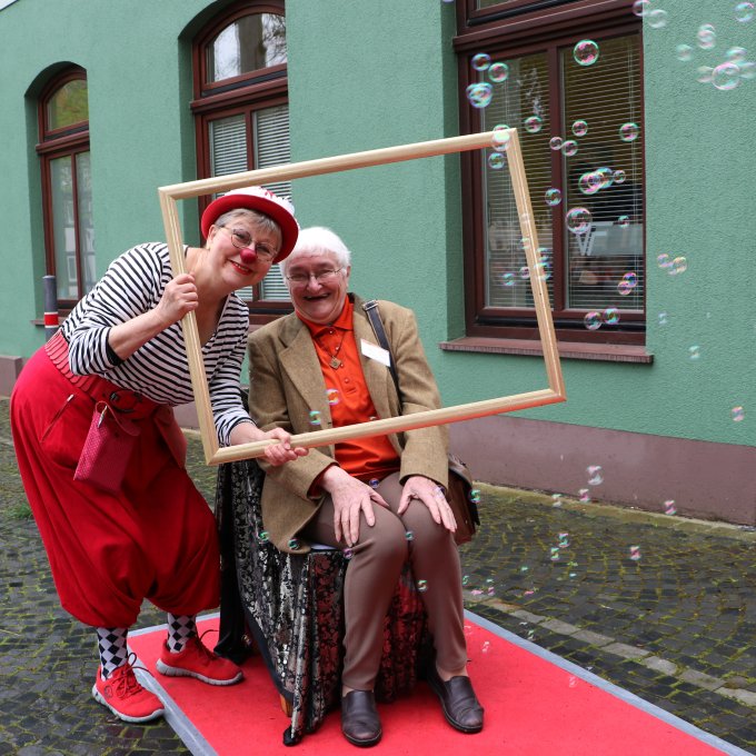 Ein Clown unterhält eine Seniorin im Rollstuhl. Im Hintergrund sind Seifenblasen zu sehen.